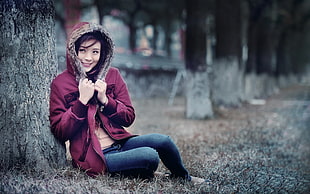 shallow focus photography of woman wearing maroon hoodie sitting beside tree during daytime