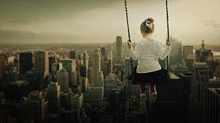 macro photography of girl sitting on swing with high rise building background