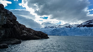 photo of body of water surrounded by black rock and mountain covered by snow under nimbostratus cloud