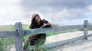 woman reading a book sitting near gray wooden fence at daytime