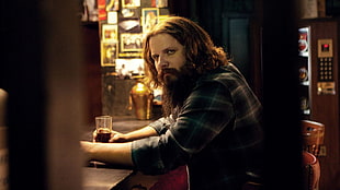 man in green plaid shirt sitting on chair while holding drinking glass