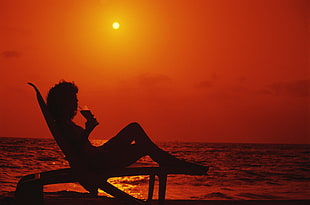 silhouette photography of woman on beach lounger holding drinking glass near seaside during sunset
