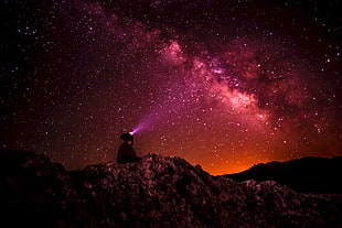silhouette of person with pink LED headlamp sits on ground overlooking sky at night time