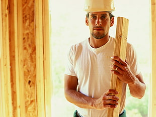 man in white crewneck shirt wearing white hard helmet holding brown wooden plank