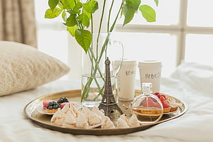 grey metal Eiffel Tower beside two disposable cups, fragrance bottle, cupcakes, and glass vase place on round brass-colored tray near window at daytime