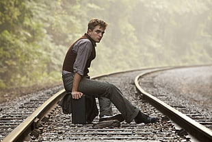 man in grey pants sitting on suitcase in train rails