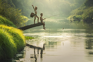 two boy's sitting on log while fishing beside river