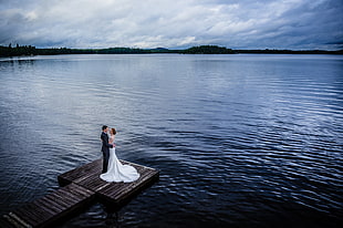 couple stands on dock near body of water under white cloud blue skies