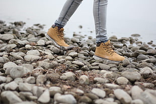 close up photo of person wearing gray jeans and brown boots walking on shore
