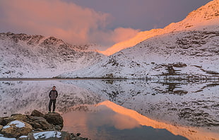 photo of man in jacket standing on big rocks beside body of water with white mountain during daytime, snowdonia