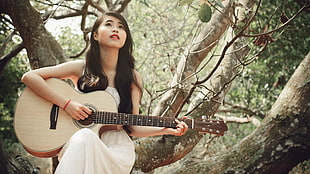 woman in white spaghetti strap dress playing white wooden cut-away acoustic guitar seated on tree branch