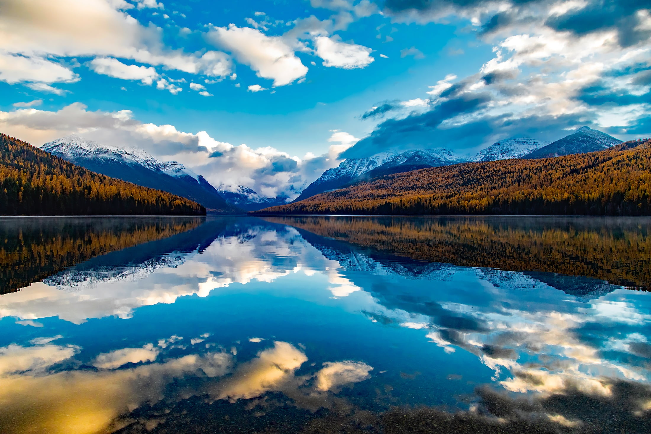 calm body of water near mountains and pine trees under white and blue sky at daytime