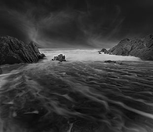 greyscale photography of body of water crashing into coastal rocks