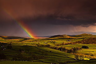 rainbow over green hill terrain under dark cloudy sky