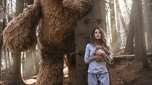 woman wearing white elbow-sleeved shirt carrying bear plush toy hiding on tree behind brown fur giant animal in forest during daytime HD wallpaper