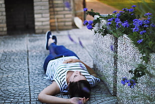 selective focus photography of a woman in white and black stripe t-shirt with blue jeans lying on grey concrete road beside purple petaled flowers