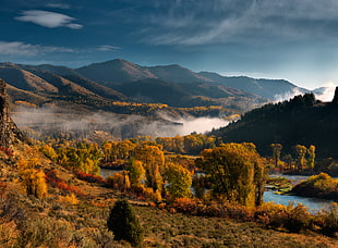 river between mountain ranges with brown trees under blue cloudy sky