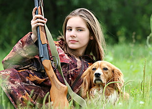 girl beside brown dog holding rifle lying on grass near trees during daytime