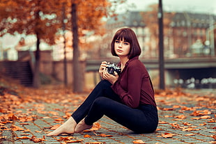 woman in maroon long-sleeved top and blue jeans sitting on ground surrounded by dried leaves tilt shift photography