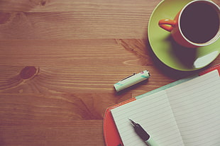 close photo of red leather covered notebook near orange ceramic mug with coffee on brown wooden table top