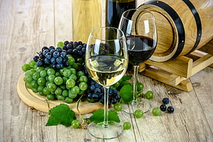 two clear wine glasses near white and red grapes and wine barrel