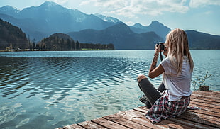 woman wearing white t-shirt sits on brown wooden platform taking photo of tree line and mountain range