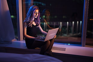 woman with black V-neck sleeveless bodycon mini dress sitting near window pane and using gray laptop computer