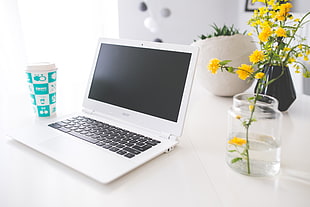 white Acer laptop beside white and blue tumbler and yellow petaled flowers in clear glass vase
