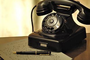 black rotary dial telephone beside black sign pen with gray paper on brown wooden surface