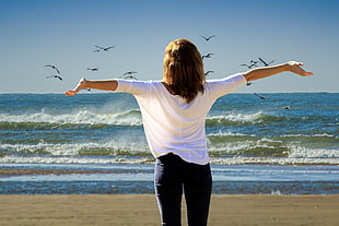 woman in white long-sleeve shirt and black fitted jeans facing wavy ocean under clear blue sky