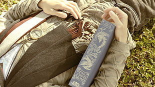 person in brown zip-up jacket holds blue labeled book lays on green grass