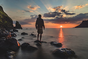 man in hoodie standing on stone beside ocean during sunset