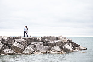 couple standing on gray rock formation on sea water under white sky