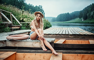 woman in white bikini top, blue shorts, and brown hat sitting on brown wooden dock