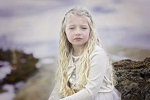 female kid wearing white cardigan sitting on brown rock