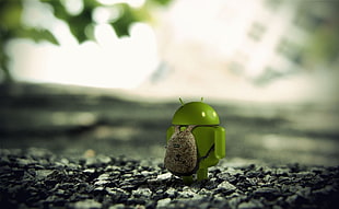 depth of field photography of green Android toy carrying bag standing on gray stones