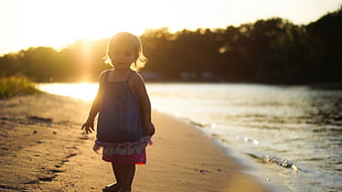 selective focus photography of girl in blue sleeveless dress stands on seashore during golden hour