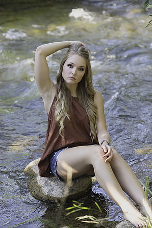 woman wearing brown sleeveless top sitting on river rock posing for photo