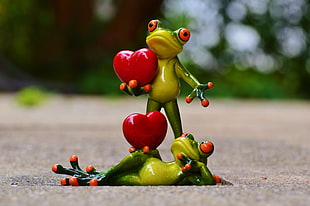 two green frog holding a heart ceramic figurine on gray road