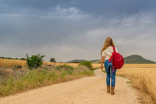 woman wearing white top and blue denim jeans with red backpack walking on rocky road near brown grass field at daytime