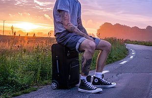 man in gray crew neck shirt sitting on soft-side luggage on gray concrete road HD wallpaper
