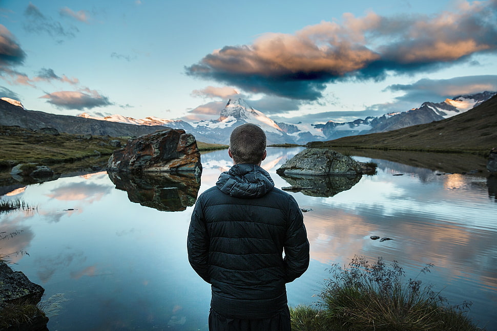 man facing the water with rocks under the gray cloudy sky HD wallpaper