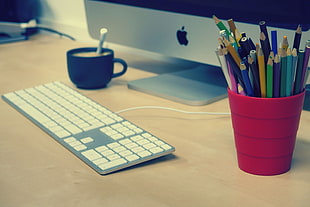 assorted color pencils in red cup near silver iMac and Mac Keyboard on beige wooden surface