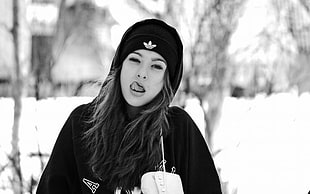 grayscale photo of woman wearing jacket and knit cap showing tongue