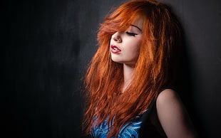woman with orange hair wearing blue tank top leaning on grey wall