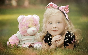 shallow focus photograph of a girl in black dress planking on green grass with her pink bear plush toy during daytime