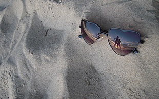 black heart-shaped sunglasses on sand reflecting the image of two persons