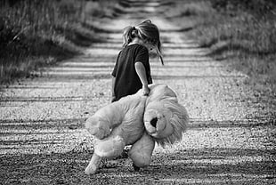 greyscale photography of girl wearing shirt standing on pathway holding life size animal plush toy