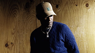 man wearing blue jacket and brown baseball cap stands beside wooden wall