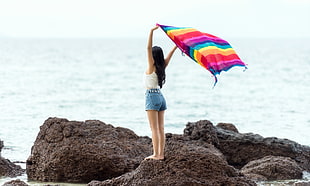 photo of woman holding shawl while standing on rock near in the sea